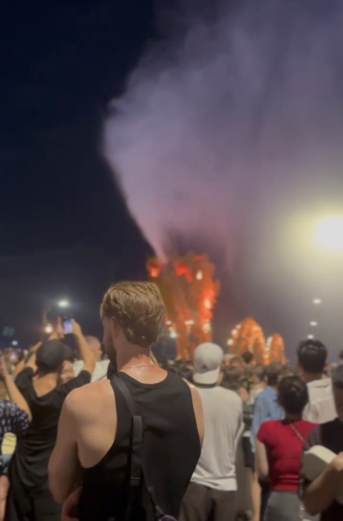 Smoke and light rising over Dragon Bridge Da Nang after the fire and water show, with crowds watching from the riverbank