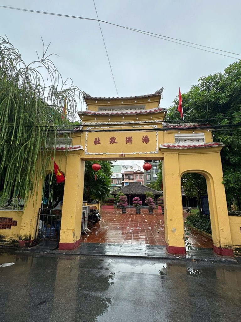 The yellow triple gate of Đình Làng Hải Châu in Da Nang, with Chinese characters, red lanterns and the communal house behind a wet tiled courtyard