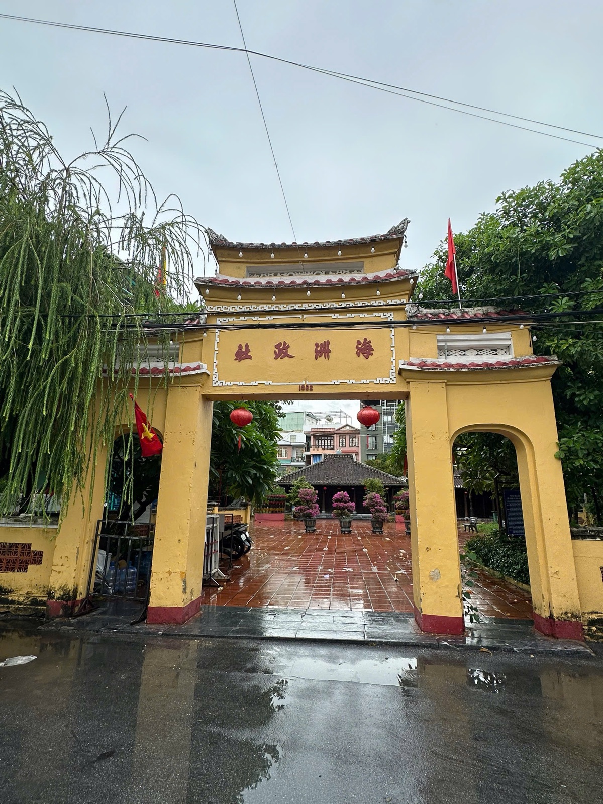 The yellow triple gate of Đình Làng Hải Châu in Da Nang, with Chinese characters, red lanterns and the communal house behind a wet tiled courtyard