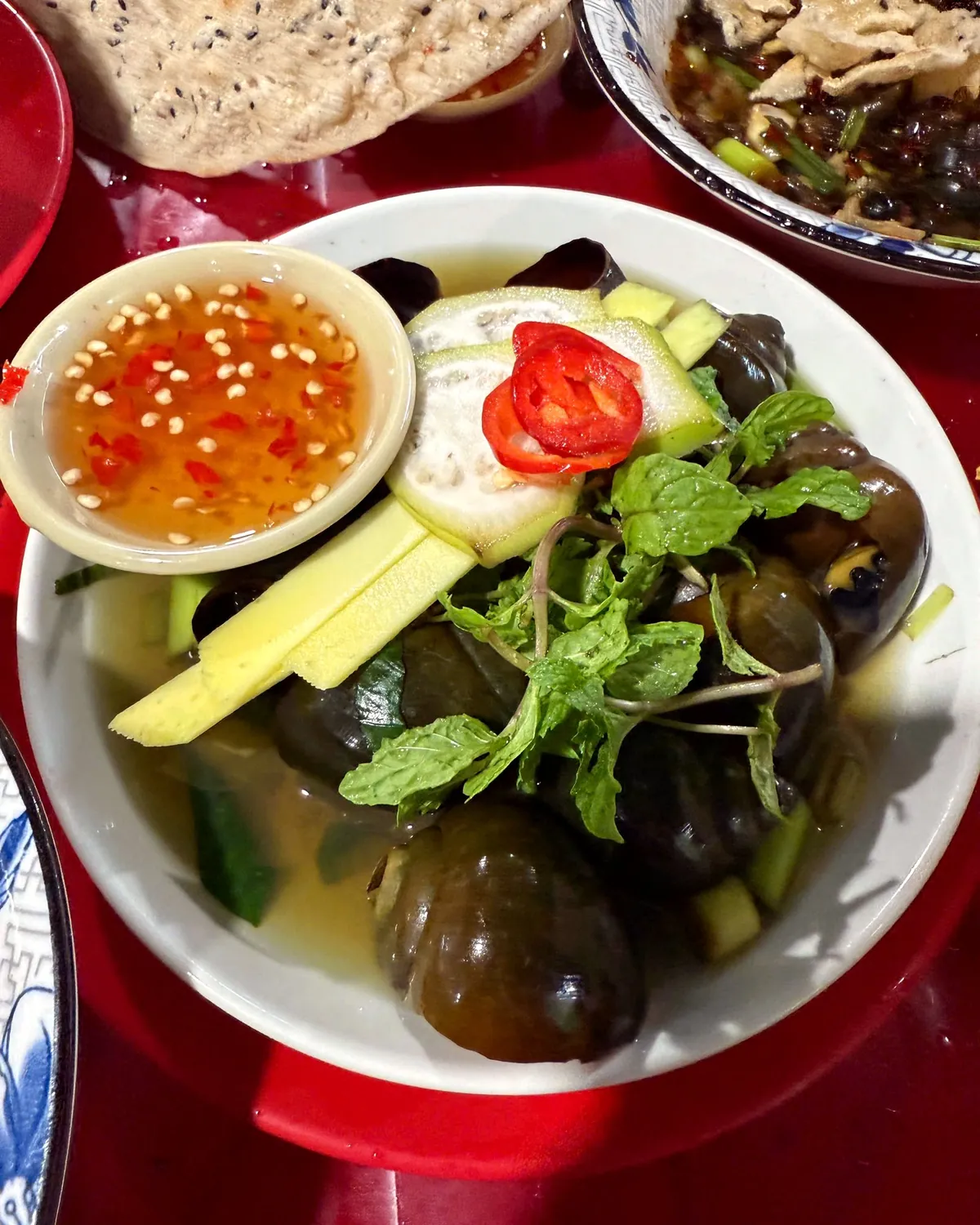A bowl of large apple snails with mint, ginger strips, cucumber and red chili, next to a small dish of ginger fish-sauce dip