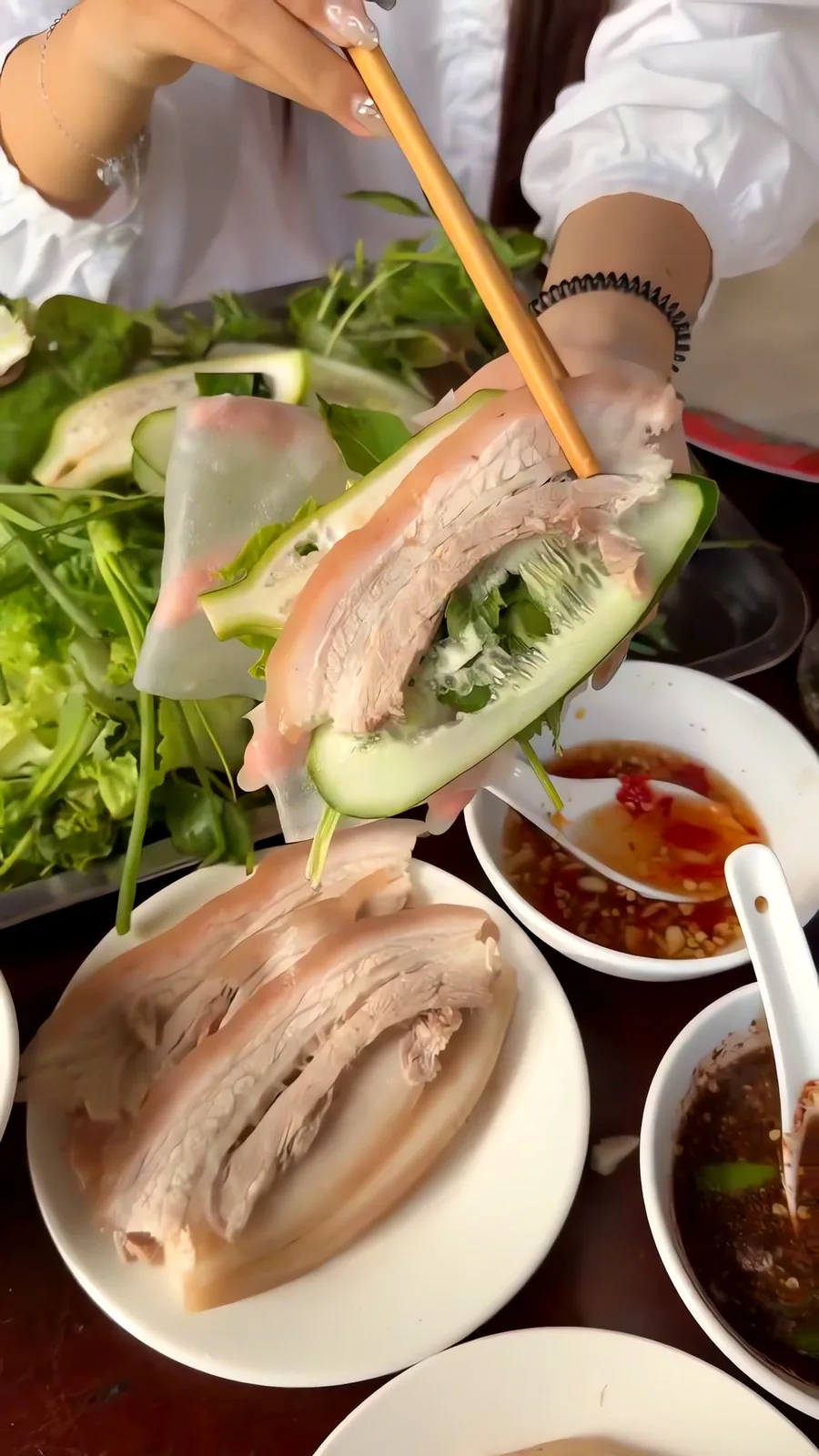 Rolling bánh tráng thịt heo at Bi Mỹ: chopsticks lay boiled pork belly on cucumber and herbs in rice paper, with mắm nêm and chili sauce below