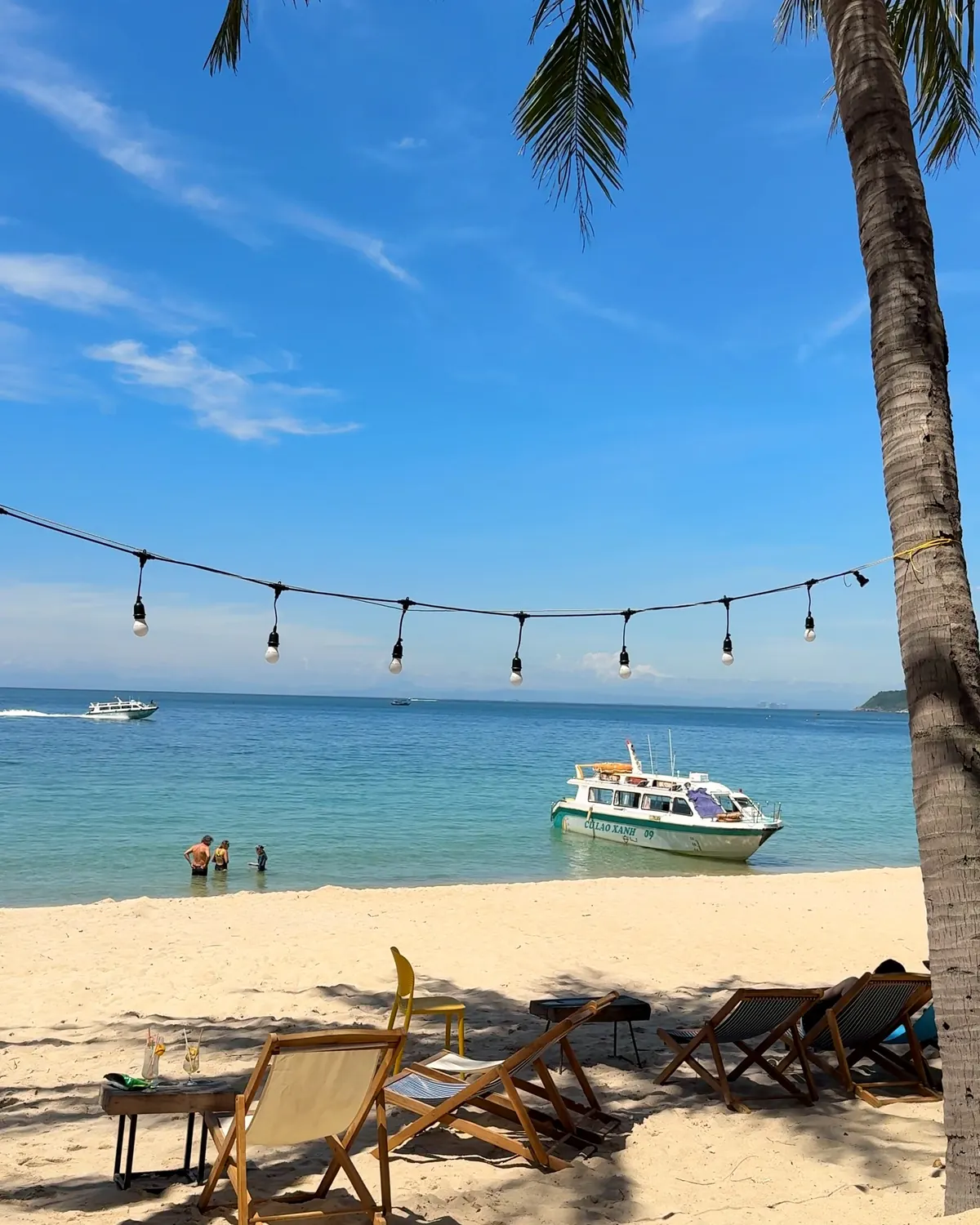 String lights hanging between palm trees over a white-sand beach and turquoise sea on Cù Lao Chàm, with a speedboat moored offshore