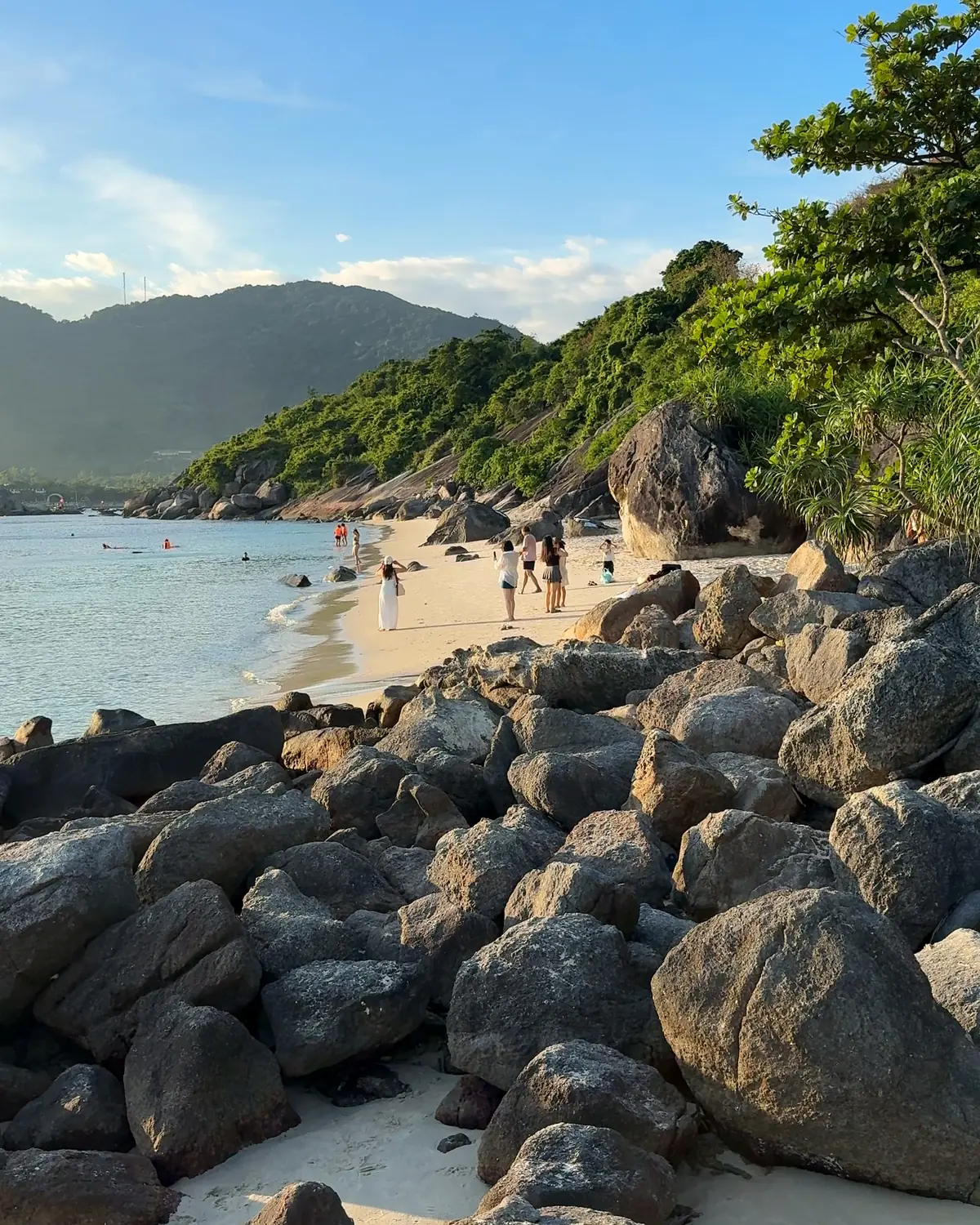 Clear water lapping granite boulders and a sandy beach on Cù Lao Chàm, with swimmers in the shallows
