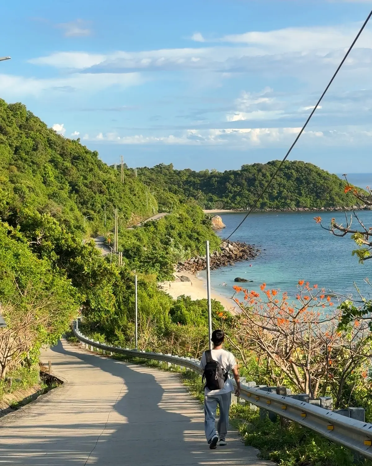 A traveller walking along the quiet coastal road on Cù Lao Chàm, with green hills, a small bay and blue sea ahead