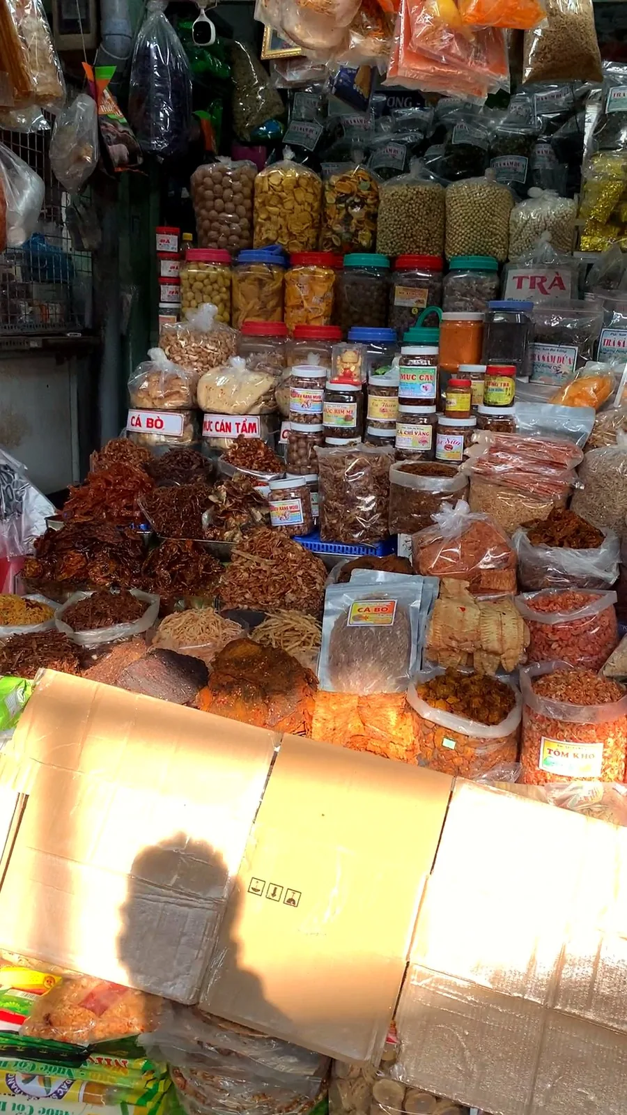 A dried seafood stall at Con Market with piles of seasoned dried squid and fish, jars of snacks and handwritten labels for cá bò and mực cán tẩm