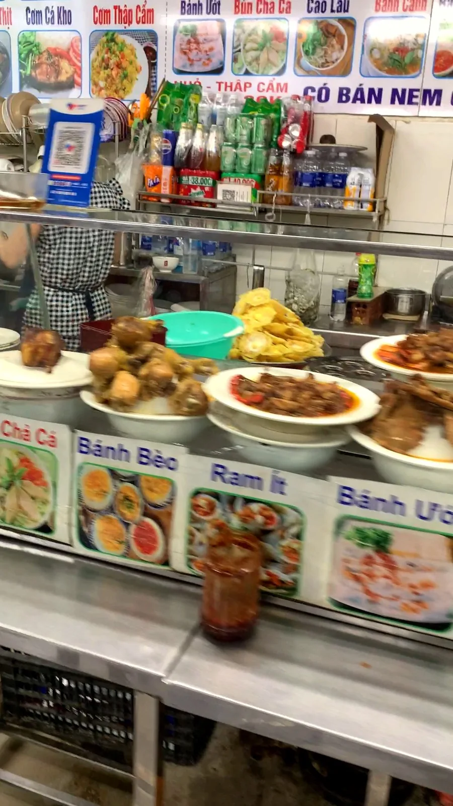 An indoor food-court stall at Con Market with bowls of food and picture menus for bánh bèo, bánh ướt, cao lầu and bún chả cá