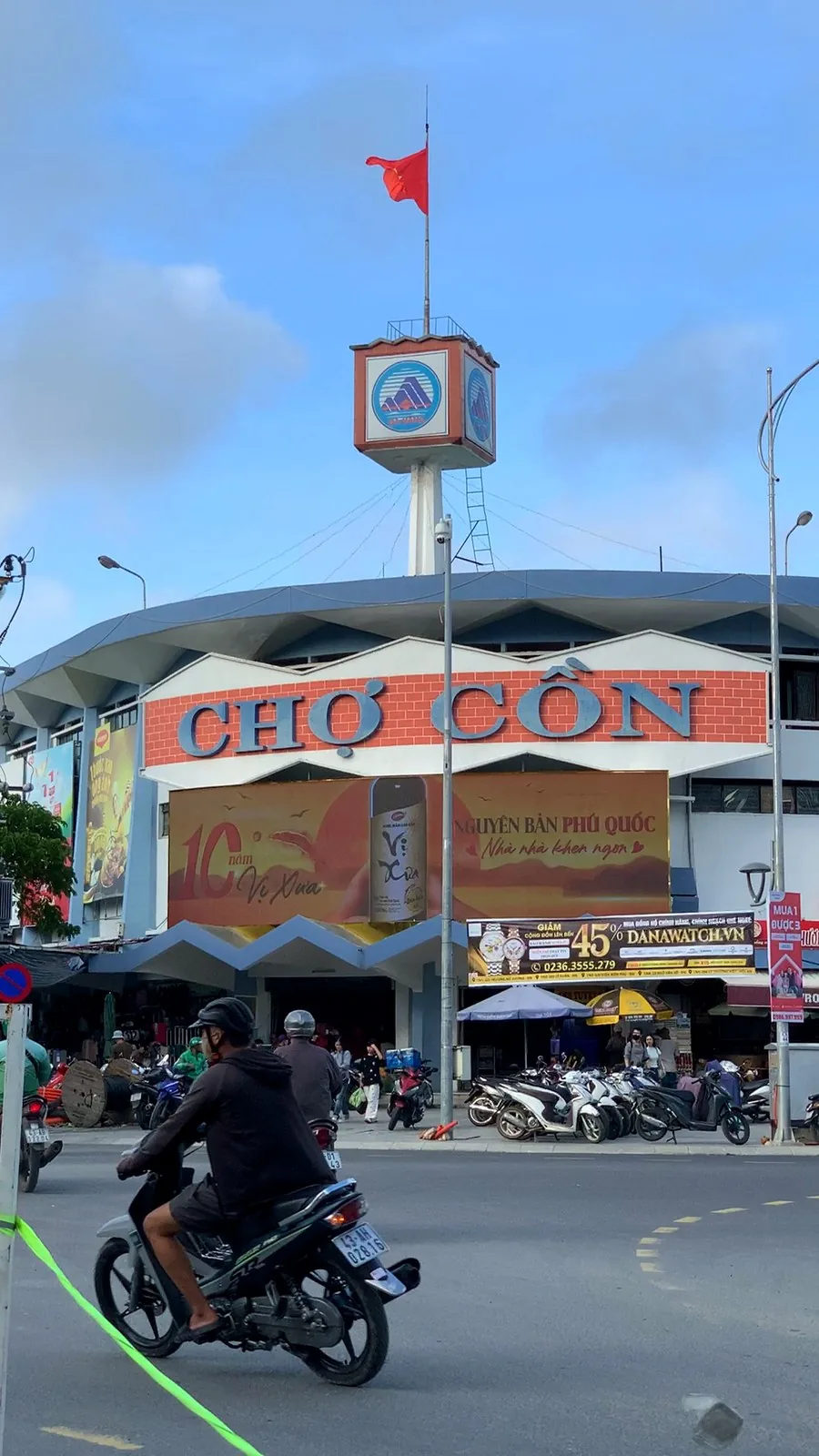 The front of Con Market market on Hùng Vương street in Da Nang, with its red "Con Market" sign, a tower with the city logo and motorbikes parked outside