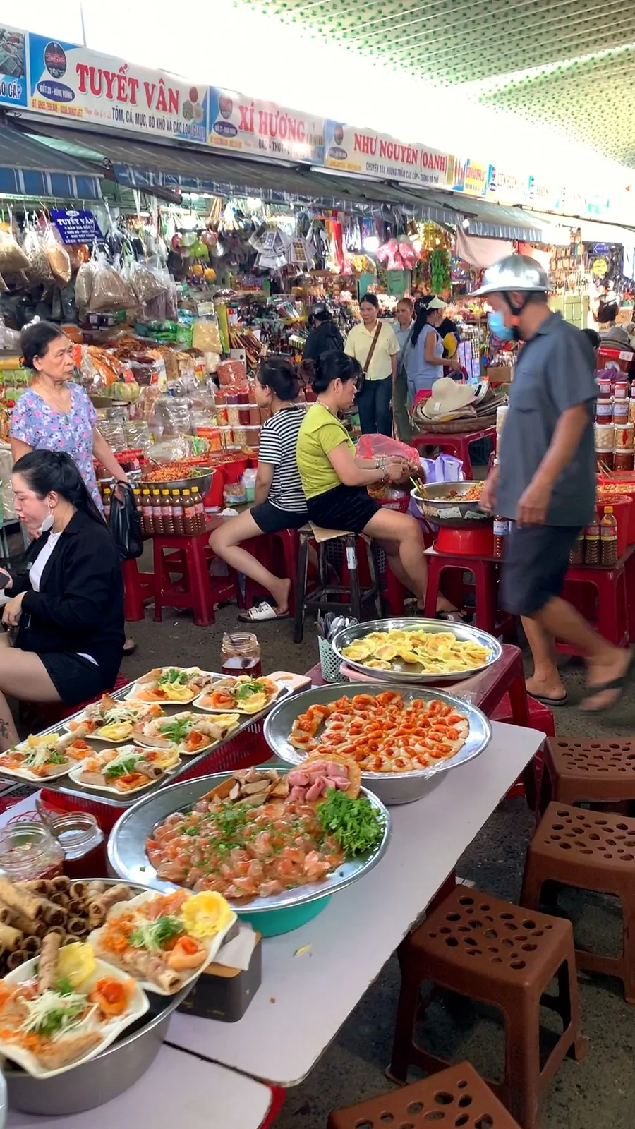 Platters of ready-made Da Nang snacks on a table with brown plastic stools in the busy food area of Con Market