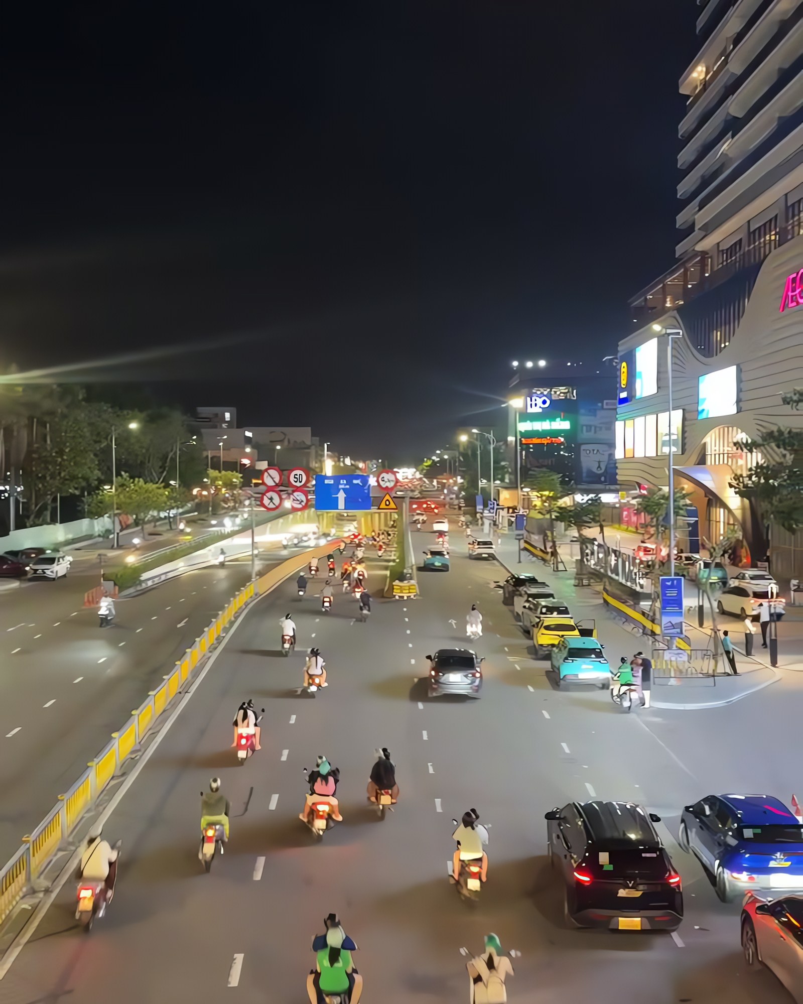 Da Nang at night seen from above, motorbikes and cars on a lit boulevard beside a shopping mall