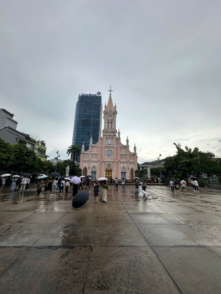 Da Nang Cathedral (Pink Church) on a rainy day, with visitors holding umbrellas in the wet courtyard