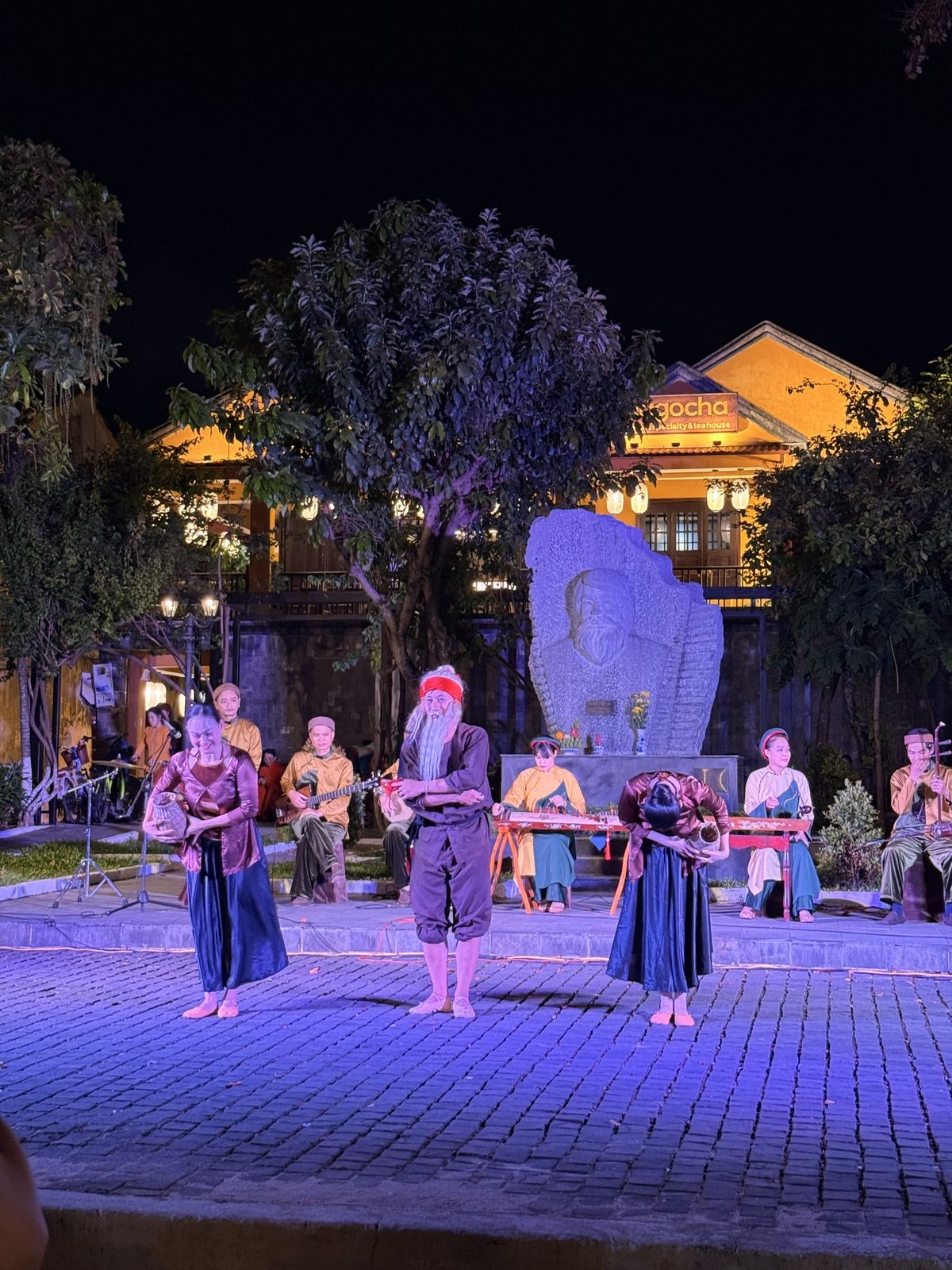 Folk arts program on stage at Kazik Park, Hoi An: a comic folk skit with an old man and two women in front of the Kazik memorial