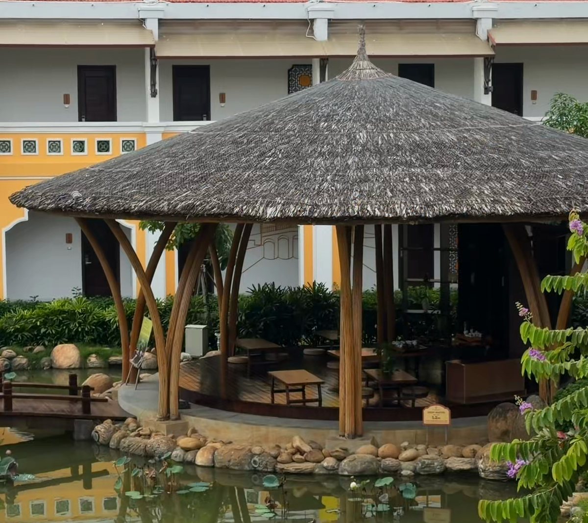 A thatched gazebo over a lily pond in the gardens of Hoi An Memories Land, with a yellow two-storey building behind