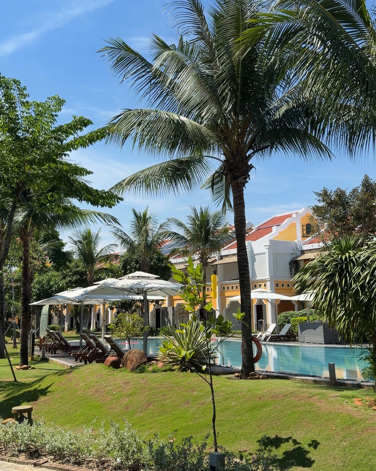 Green lawn and palm trees in the garden of Hoi An Memories Land, with yellow villas and white umbrellas by the pool in the distance