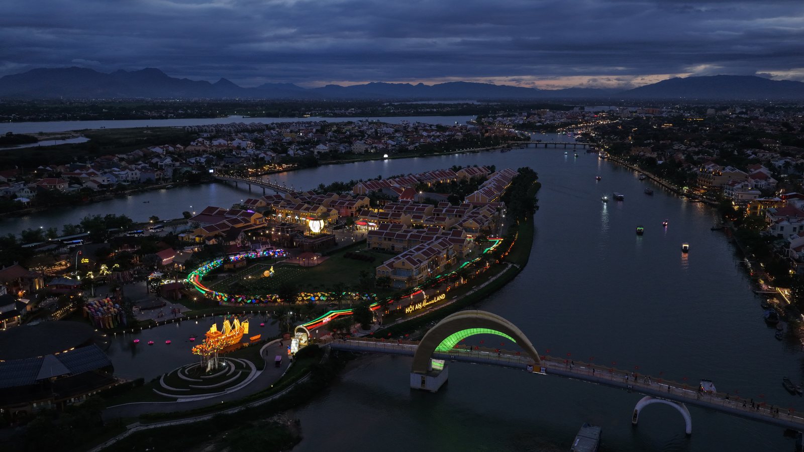Aerial view at dusk of Hoi An Memories Land lit up on its island in the Hoài River, with Hội An Old Town and the mountains behind