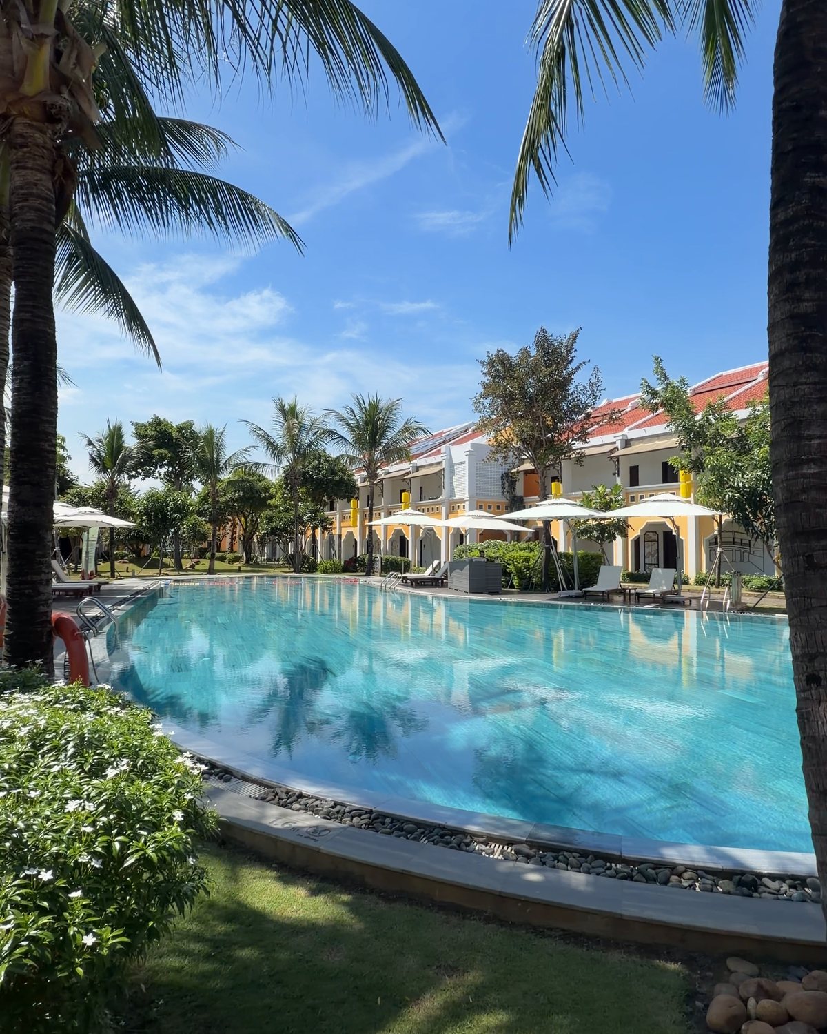 Turquoise swimming pool at Hoi An Memories Land with palm trunks in the foreground, white umbrellas and two-storey villas with red roofs behind