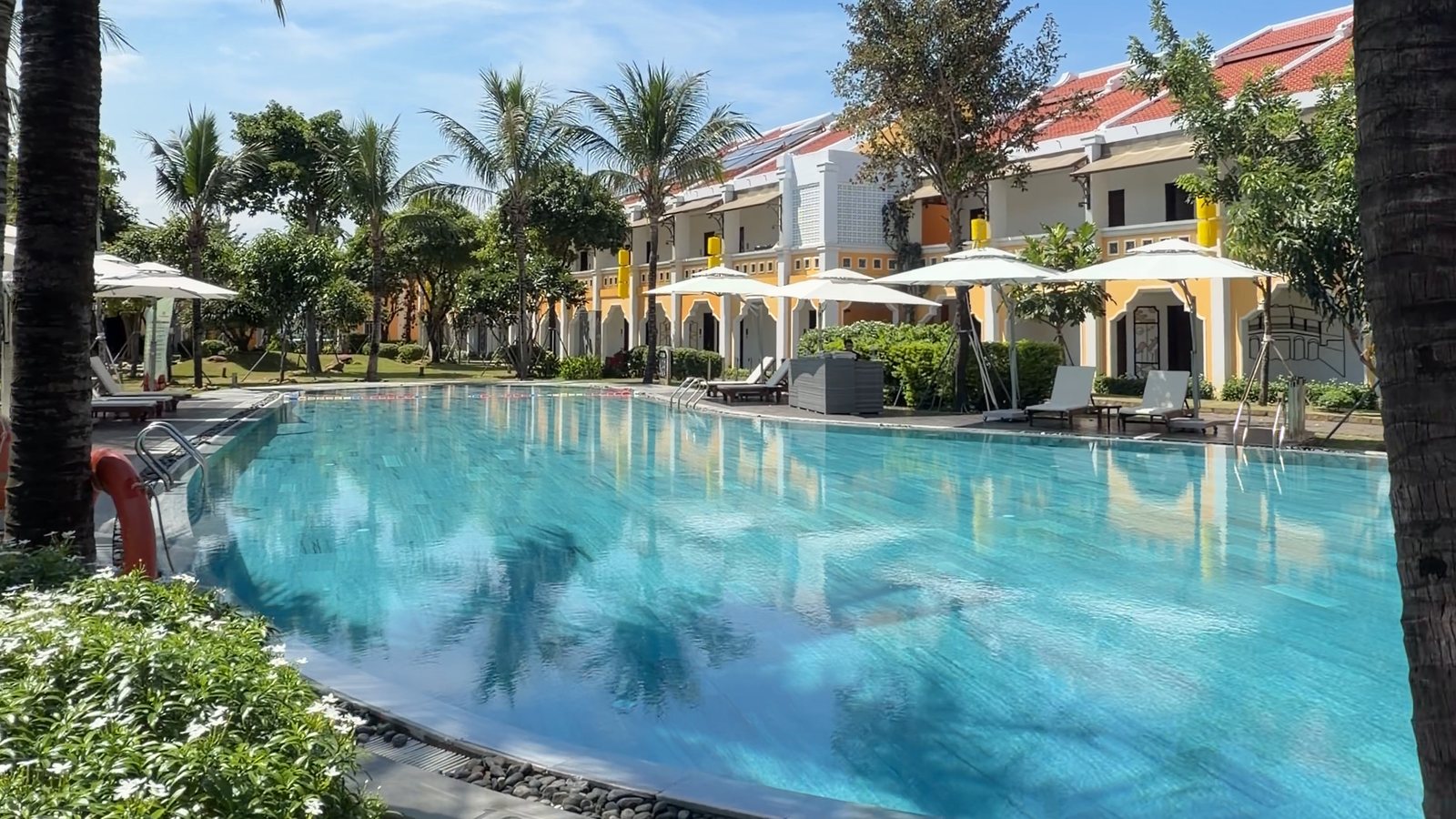 The pool at Hoi An Memories Land resort, framed by tall palm trees, with white umbrellas and yellow villas behind it under a blue sky