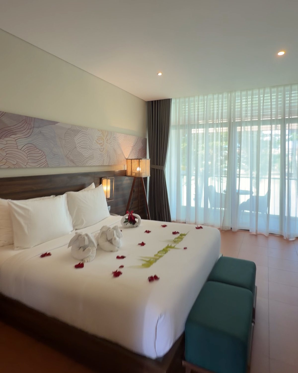 A guest room at Hoi An Memories Land: a large white bed decorated with towel swans and flower petals, teal footstools and floor-to-ceiling curtained windows