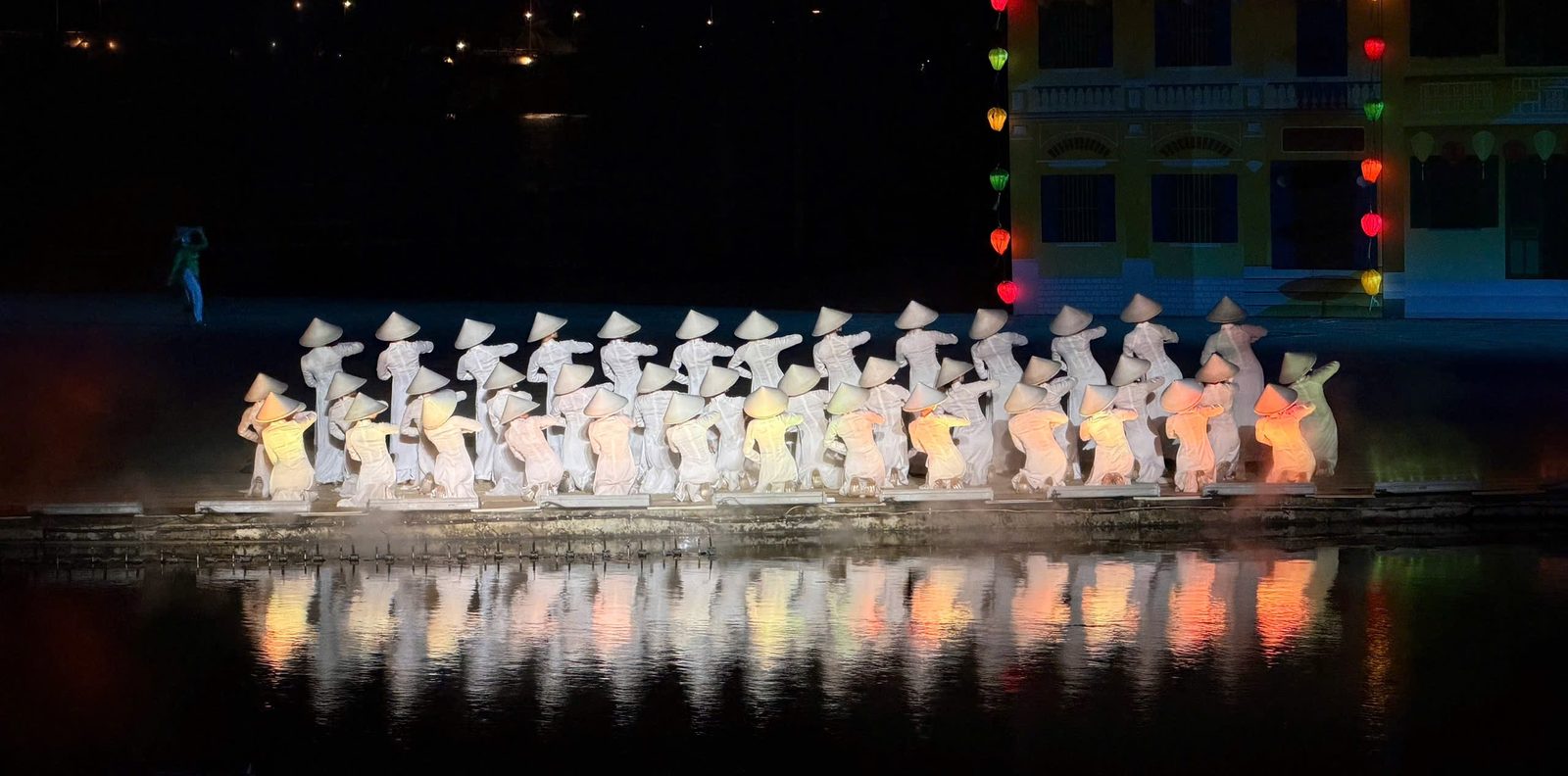 Rows of performers in white áo dài and conical hats kneel on the river stage, their reflections glowing in the water at the Hoi An Memories show