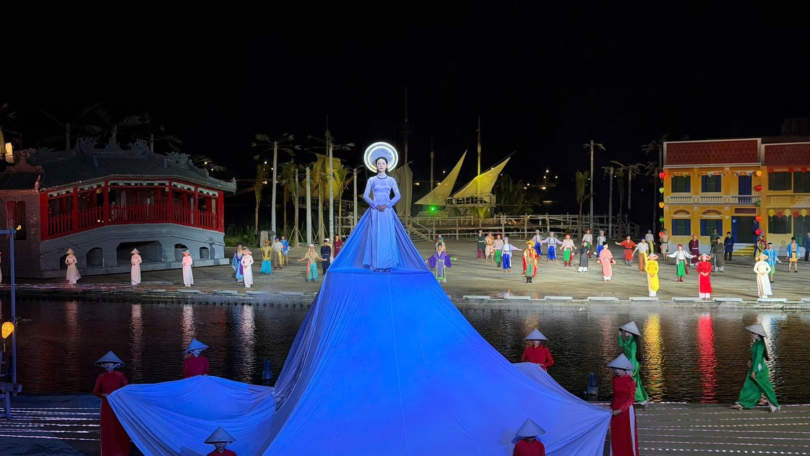 Hoi An Memories show finale: a woman in a white áo dài with a glowing halo headpiece stands on a huge blue-lit train on the river stage, with performers in áo dài and conical hats around her