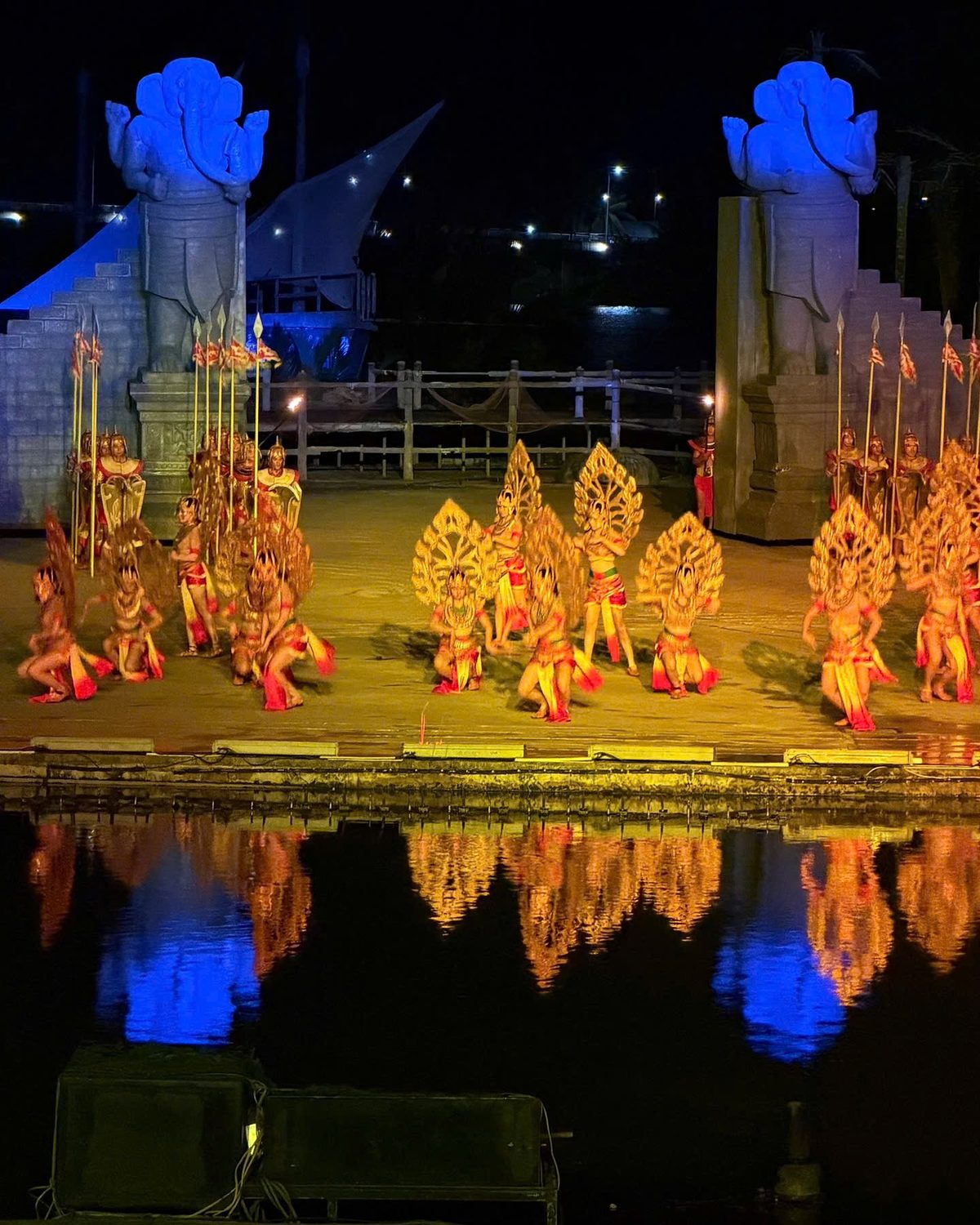 Cham dancers in golden leaf-shaped headdresses perform between two large blue Cham statues, reflected in the river at the Hoi An Memories show