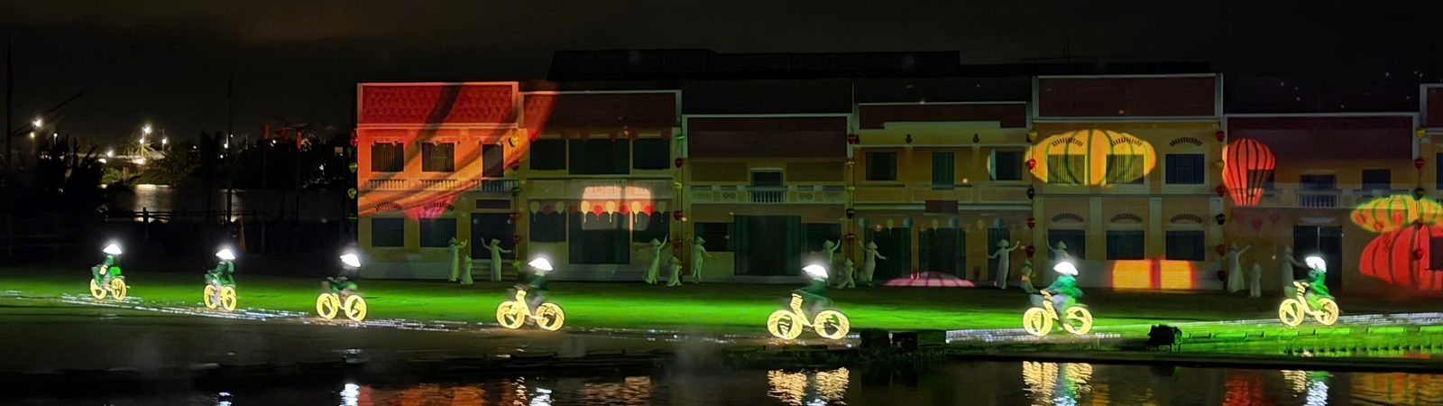 Women in áo dài ride bicycles with glowing light-outline frames along a green-lit path in front of the old-town facades in the Hoi An Memories finale