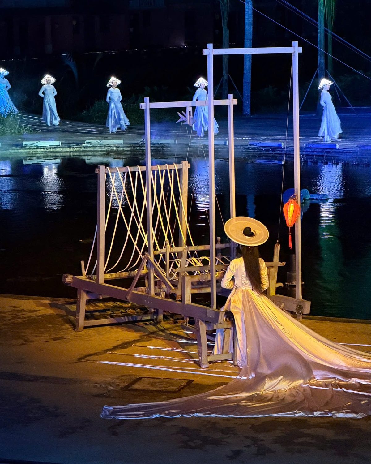 Opening act of the Hoi An Memories show: a woman in a conical hat and a long silver train sits at a wooden silk loom on the riverbank, with women in white áo dài walking along the water behind her