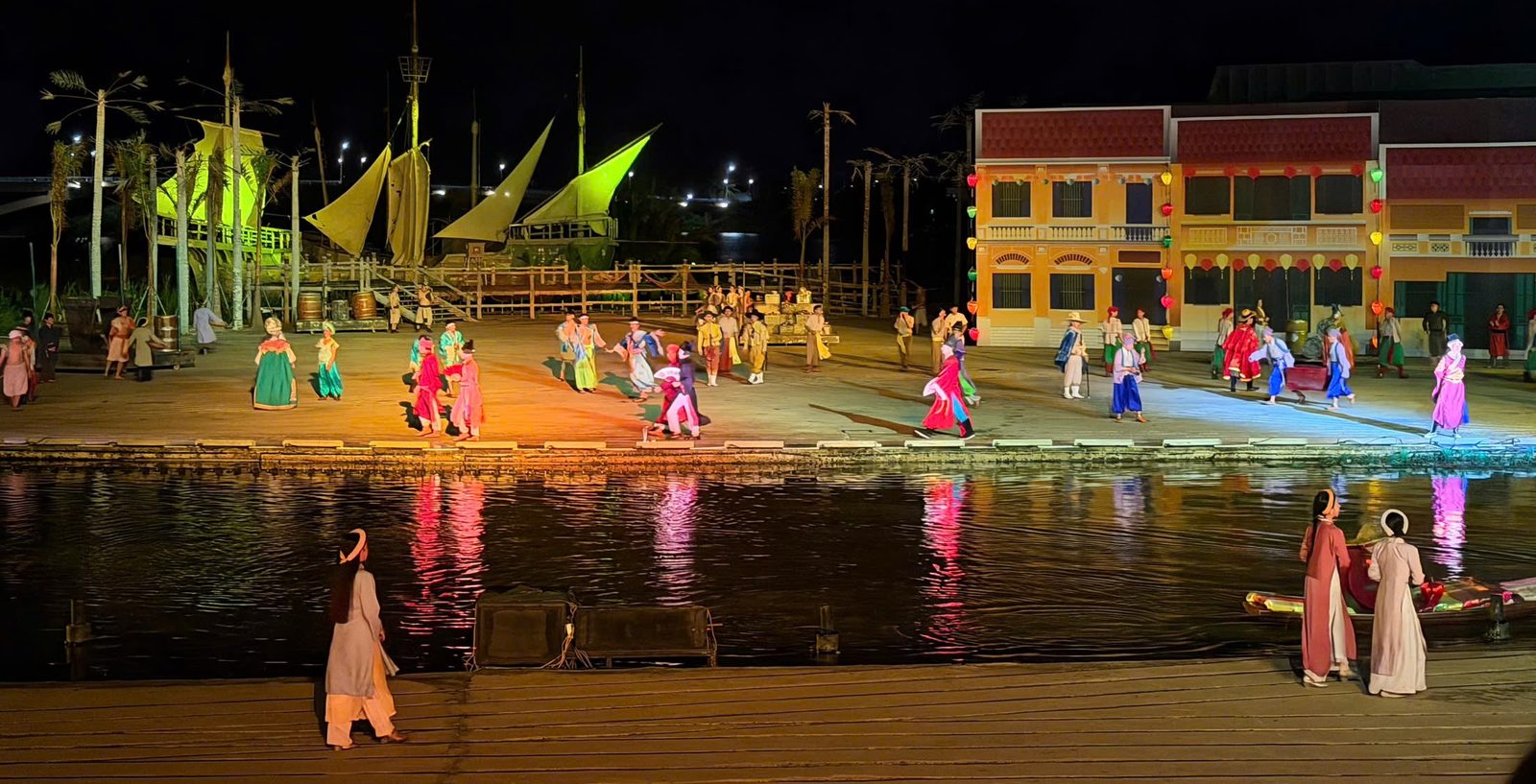 The trading port act of Hoi An Memories: performers in colourful costumes fill a riverside market scene with yellow-sailed ships and Hoi An shophouses behind
