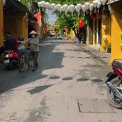 A woman in a conical hat walks her bicycle under silk lanterns on a nearly empty street in Hoi An Old Town on a sunny October morning
