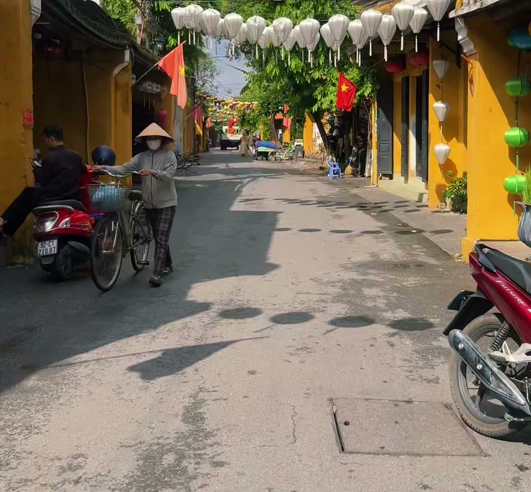 A woman in a conical hat walks her bicycle under silk lanterns on a nearly empty street in Hoi An Old Town on a sunny October morning