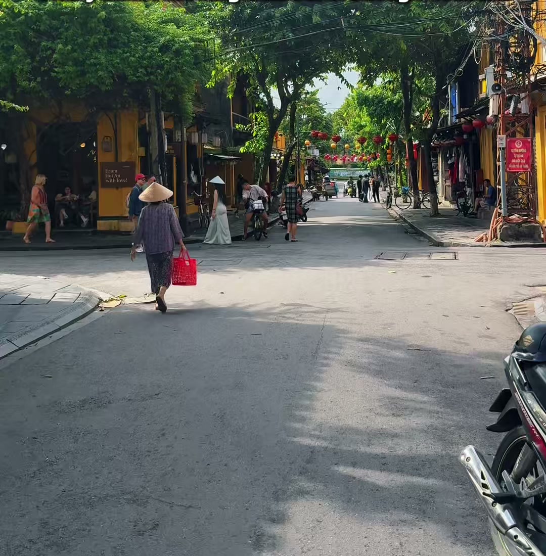 A woman in a conical hat carries a red bag across a sunny, quiet street in Hoi An Old Town on 20 October 2026