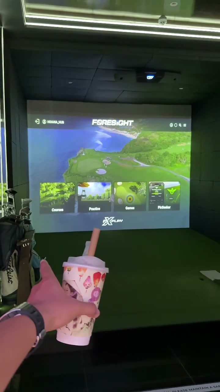Virtual golf simulator screen showing a golf course, with a drink in hand, at Hoiana Resort & Golf