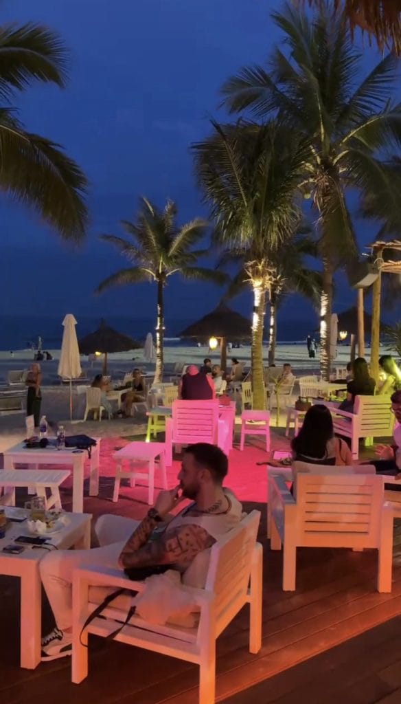 The lit-up beachfront terrace at The Holiday Beach Club & Dining on My Khe Beach at blue hour, with palm trees and white lounge chairs