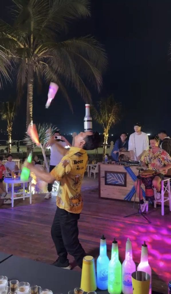 A flair bartender juggling glowing bottles beside the DJ and a drummer at The Holiday Beach Club & Dining, Da Nang