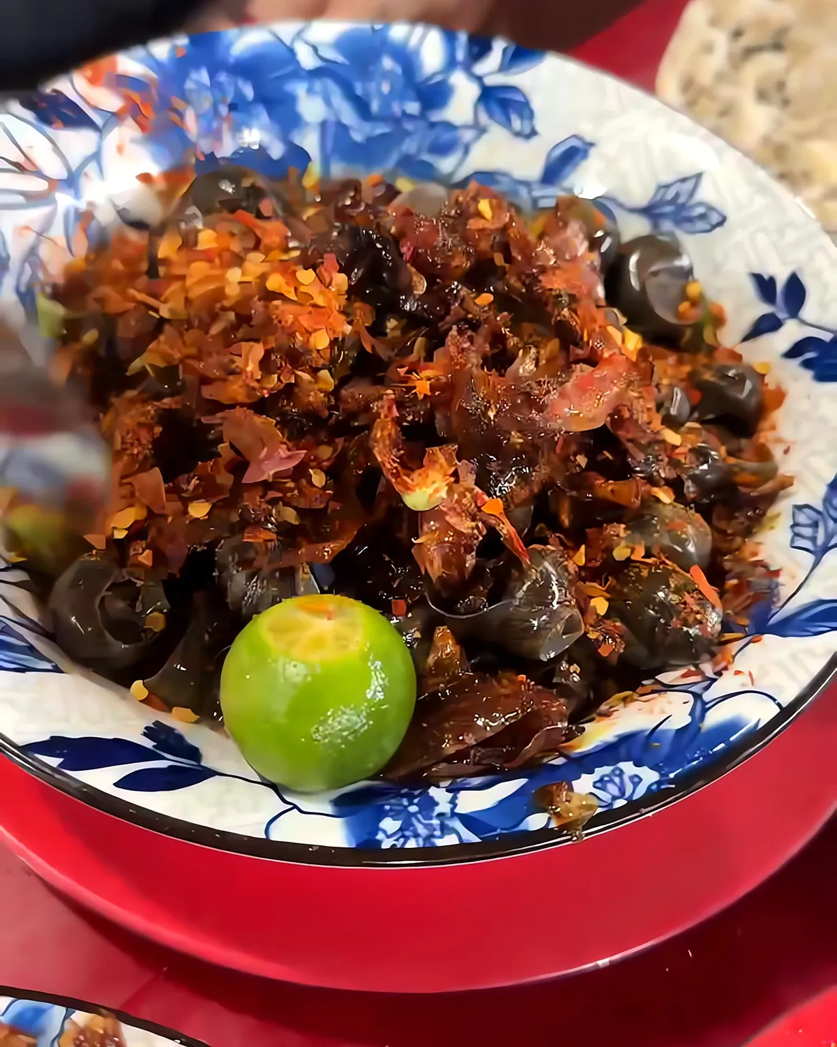 A spoonful of chili flakes over a bowl of glossy ốc hút snails and fried shallots, with a kumquat