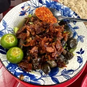 A blue-and-white bowl of ốc hút snails topped with fried shallots, with kumquat halves, chili flakes and grilled sesame rice paper on a red table in Da Nang