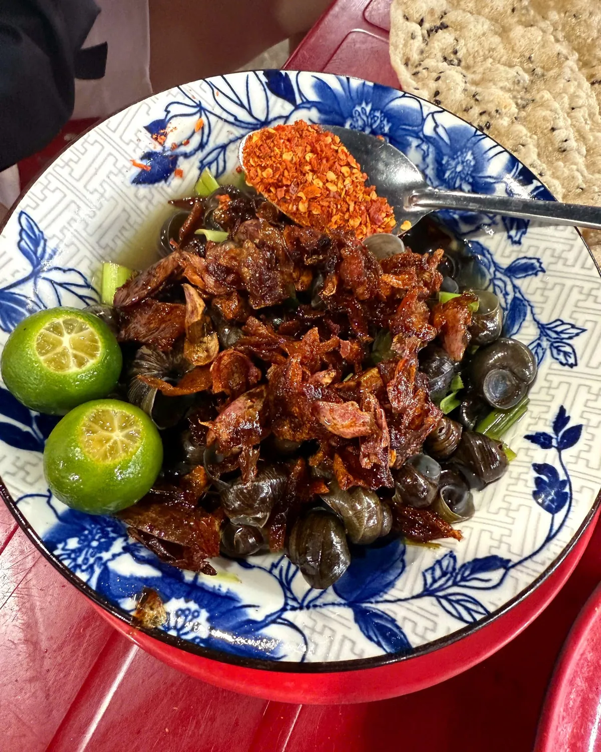 A blue-and-white bowl of ốc hút snails topped with fried shallots, with kumquat halves, chili flakes and grilled sesame rice paper on a red table in Da Nang