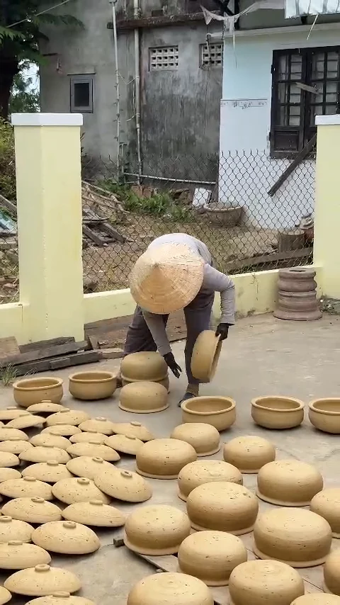 Local potter in a conical hat arranging clay pots to dry in a courtyard, Thanh Ha Pottery Village